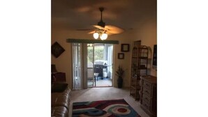 A living room with a large, colorful rug, a ceiling fan, and a view through a glass door to the outside.