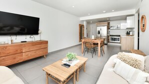 A modern living room with a flat-screen TV, wooden furniture, a dining area with wicker chairs, and a kitchen with white cabinets.