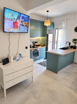 A modern kitchen with teal cabinetry, a central island, a built-in oven, and a flat-screen TV mounted on the wall. - Jacuzzi Penthouse at Gazi nights (Athina)