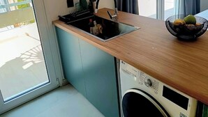 A compact kitchen with a built-in washing machine, a sink, and a small countertop with a fruit bowl. - Jacuzzi Penthouse at Gazi nights (Athina)