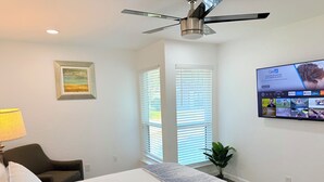 Habitación de hotel con cama, ventilador de techo, televisor montado en la pared, ventana con persianas y una planta pequeña.