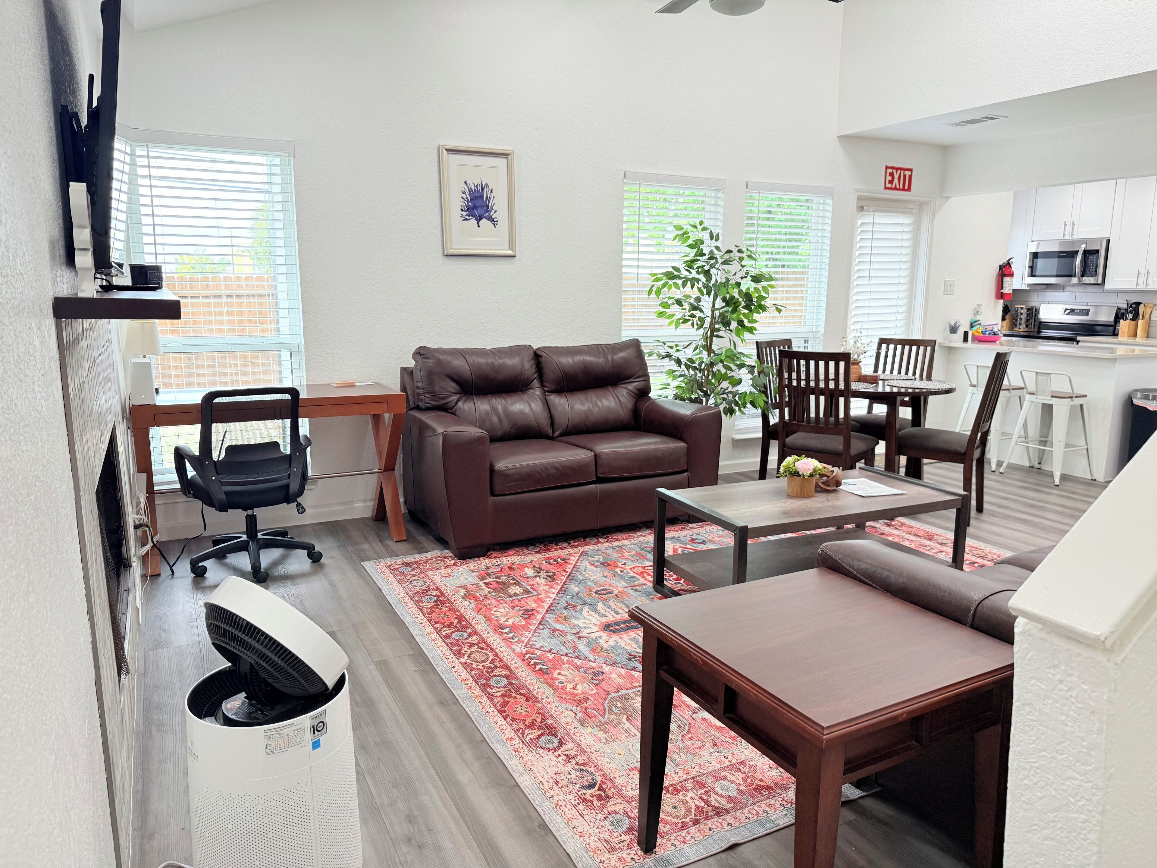 A modern living room with a brown leather sofa, a wooden coffee table, and a red patterned rug. There is a dining area with a small table and chairs, and a kitchen area with white cabinets and a microwave.