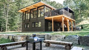 Eine moderne, zweistöckige Holzhütte mit großen Fenstern, einer Holzterrasse und einem Feuerstellenbereich aus Stein.