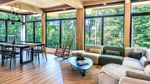 Ein geräumiger Wohnbereich mit Esstisch, Stühlen, einer Couch und einem Schaukelstuhl. Große Fenster bieten Blick auf den umliegenden Wald.