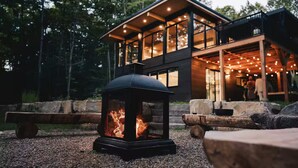 Ein modernes Haus mit einer großen Terrasse, einem Feuerbereich im Vordergrund und einem bewaldeten Hintergrund.