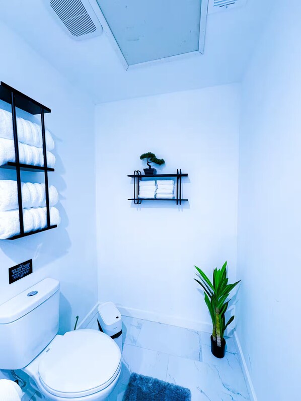 A bathroom with a toilet, a shelf with towels, a small plant, and a sign on the wall.