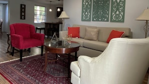 A living room with a red armchair, a beige sofa, and a coffee table with decorative items. There are two wall-mounted lamps and a decorative wall panel. - Family Getaway Near Beaches, Sleeps 8 (New Port Richey)