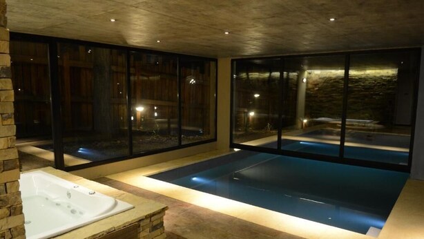 Un área de spa moderna con una bañera de hidromasaje, una piscina grande con paredes de vidrio y una pared de piedra.