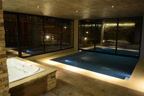 Un'area spa moderna con una vasca idromassaggio, una grande piscina in vetro e una parete in pietra.