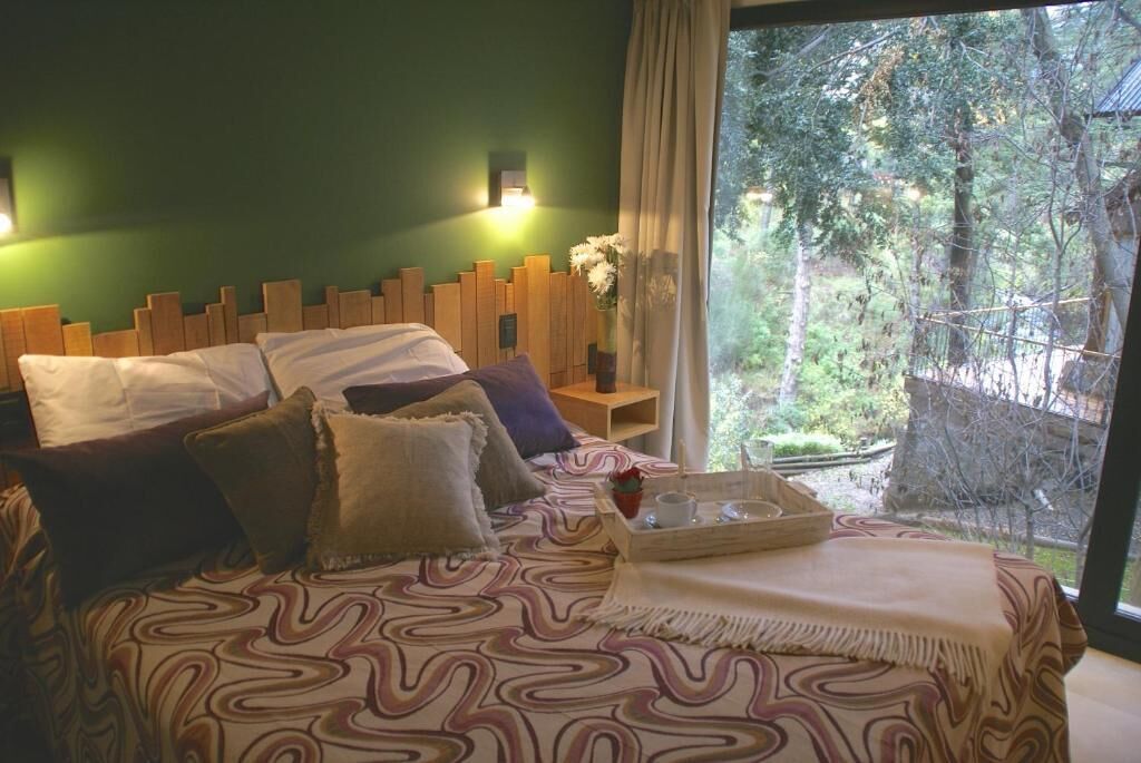 Un dormitorio con cama, almohadas, mesita de noche con lámpara y ventana con vista a árboles.