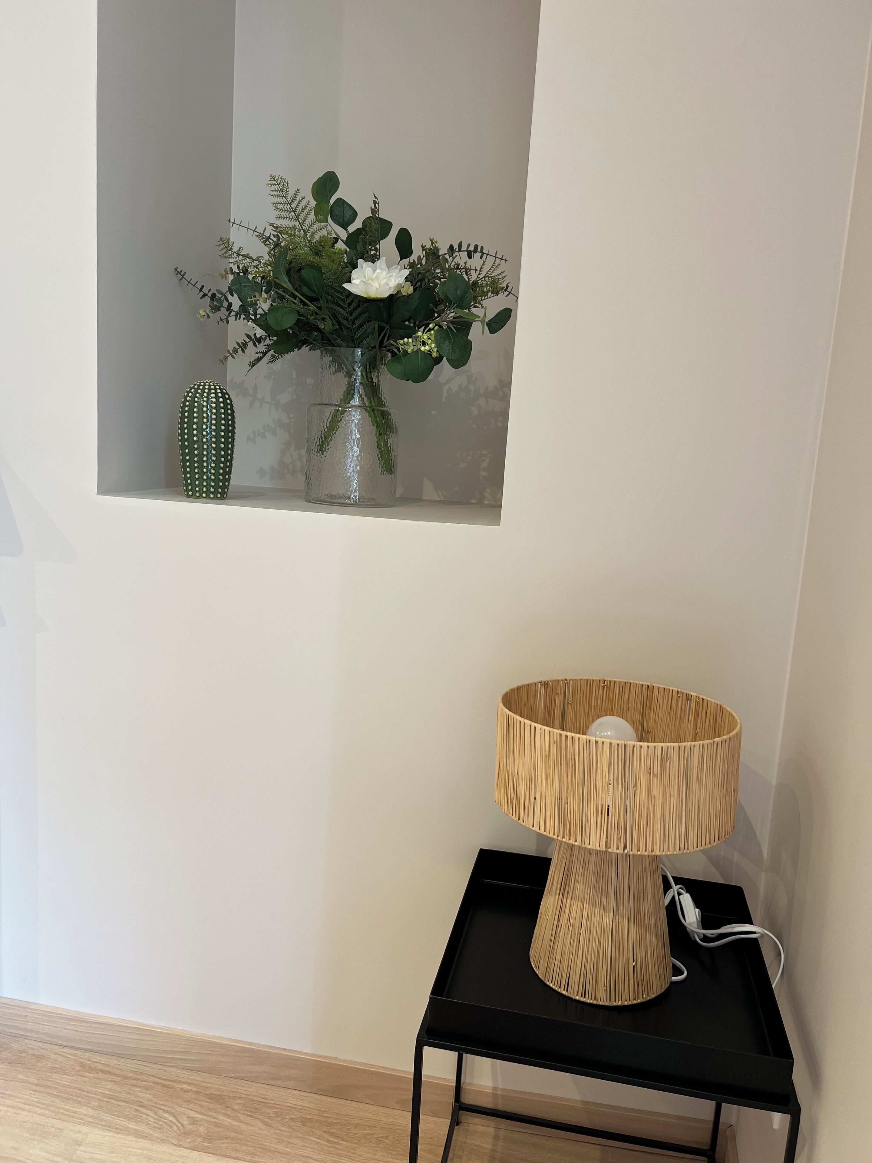 Une chambre moderne avec une table en métal noir, une lampe en fibres tressées et une petite fenêtre ornée d’un cactus et de fleurs.
