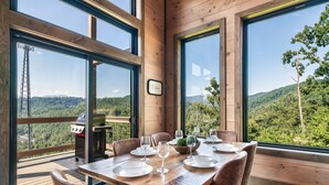 A dining area with a wooden table set for six, large windows offering a view of a forested landscape, and a grill outside.