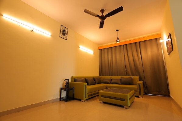 A living room with a sofa, ottoman, ceiling fan, and wall-mounted lights. - BL Homestay & Suite (Agra)