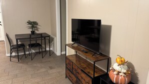A living room with a TV on a wooden stand, a dining table with chairs, and a potted plant.