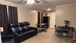 A living room with a black leather sofa, a dining table with chairs, a ceiling fan, and a potted plant.
