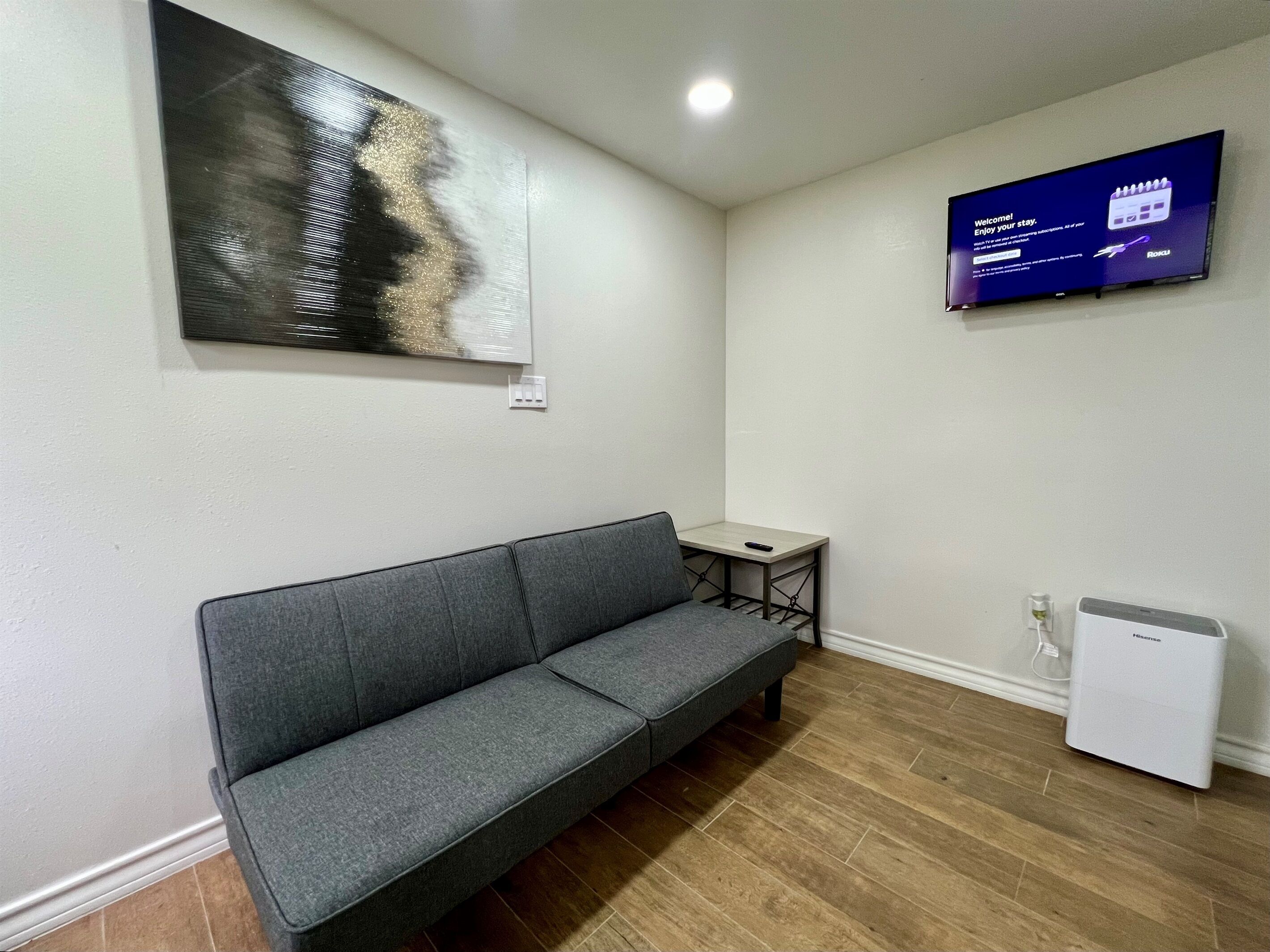 Et værelse med en grå sofa, et lille bord, et fladskærms-tv og et hvidt minikøleskab.