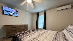 Un dormitorio con cama, cómoda de madera, ventilador de techo y televisor con pantalla mostrando una interfaz de servicio de streaming.