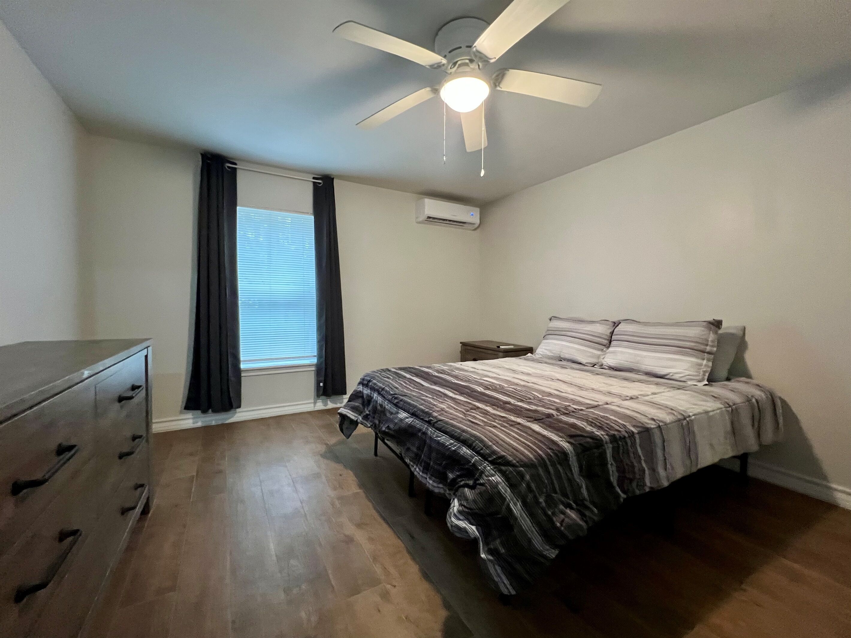 A bedroom with a bed, a dresser, a ceiling fan, a window with curtains, and an air conditioning unit.