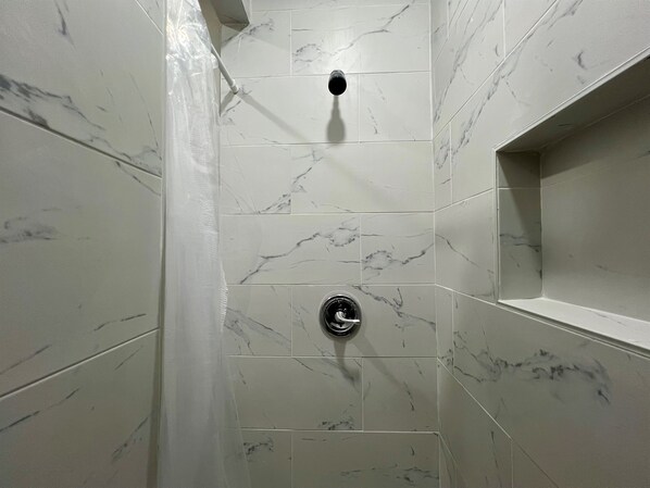 A bathroom with white marble tiles, a shower with a handheld showerhead, and a built-in shelf.