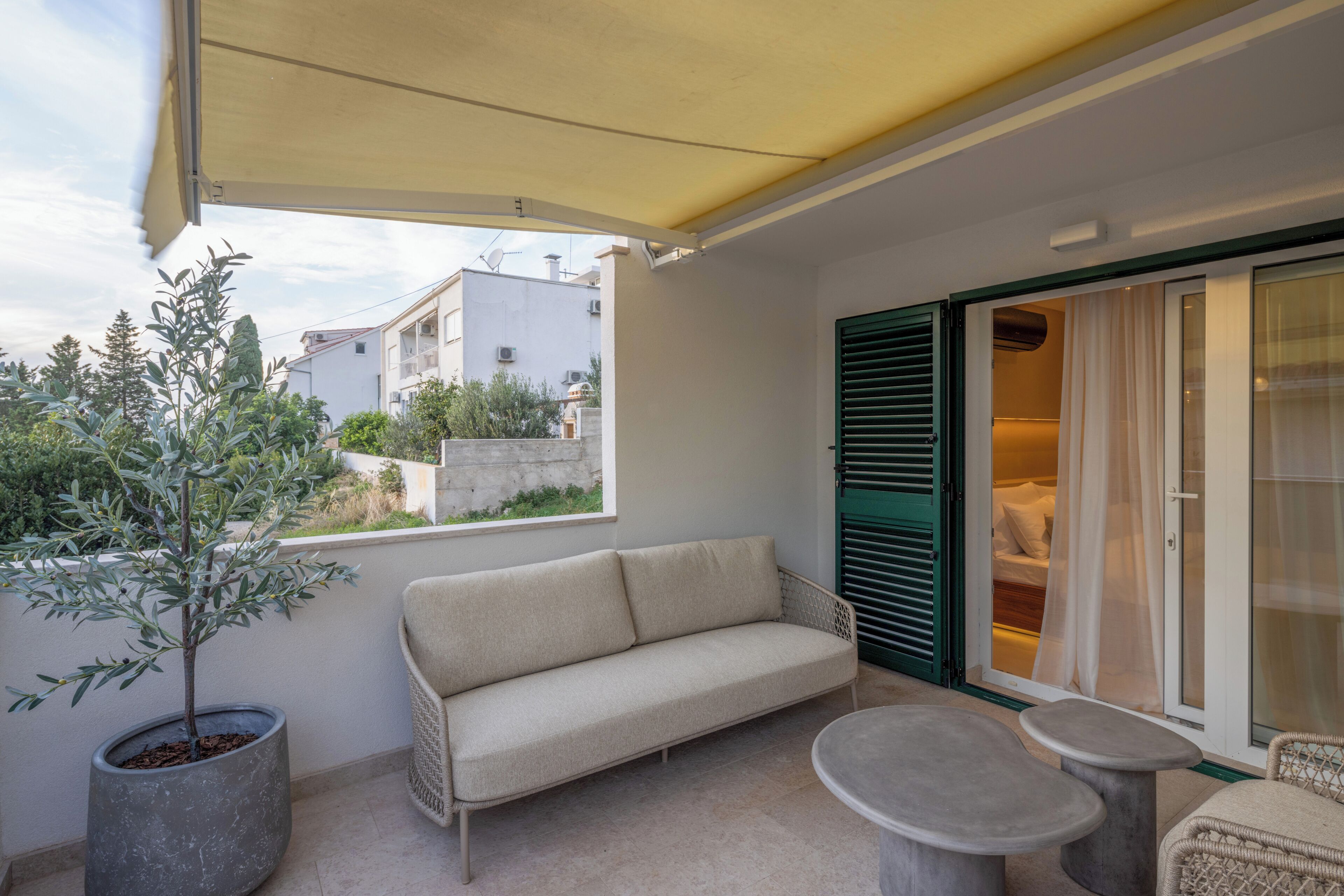 A balcony with a sofa, two small tables, a potted plant, and a sliding glass door leading to a bedroom.