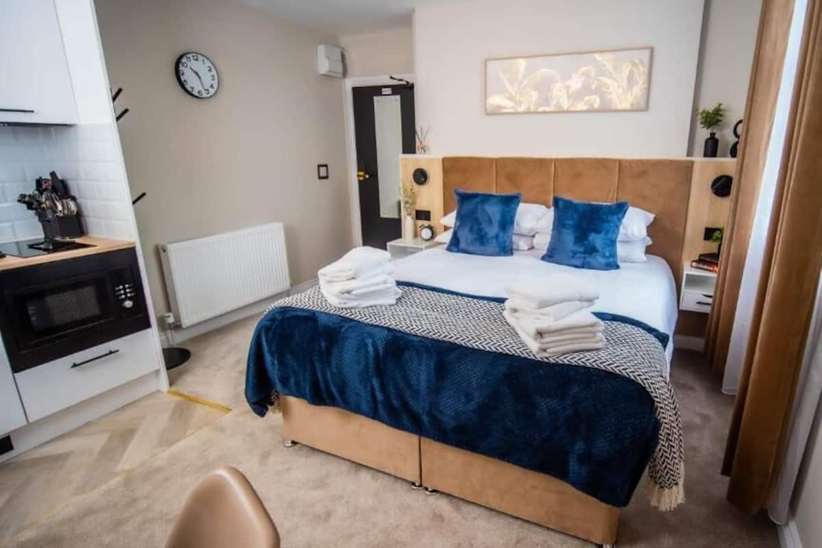 Habitación de hotel moderna con una cama grande, una mesita de noche, un reloj en la pared y vistas al pasillo.