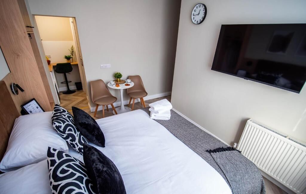 A hotel room with a bed, a TV, a clock, a small table with chairs, and a radiator.