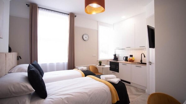 A hotel room with two beds, a small kitchenette, a clock on the wall, and a window with curtains.