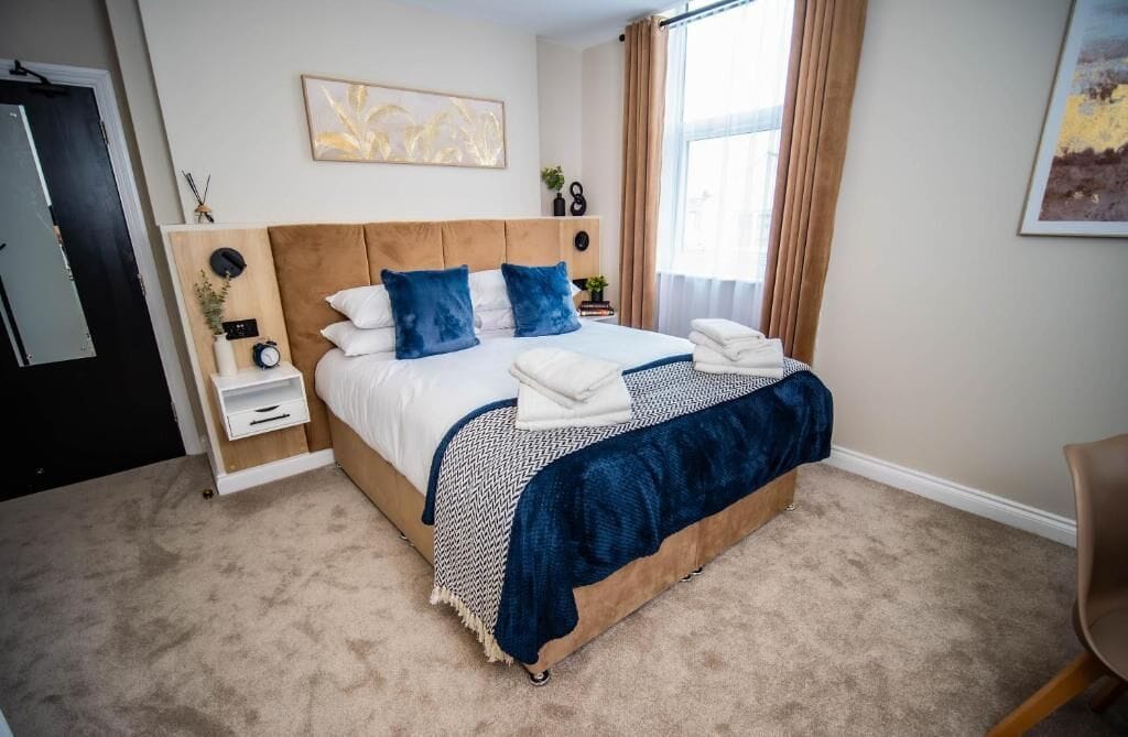 Une chambre à coucher comprenant un lit, des tables de chevet, une commode, une horloge murale, un tableau encadré et une fenêtre avec des rideaux.