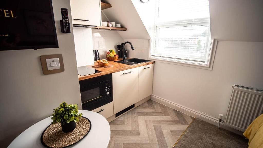 Una cocina compacta con encimera de madera, un lavamanos y un horno empotrado. Hay una pequeña mesa con una planta y una silla blanca. La habitación tiene un techo inclinado y una ventana con persianas.