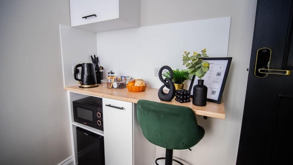 A compact kitchenette with a microwave, coffee maker, and a small oven. A green chair is positioned in front of a white cabinet, and a small plant is placed on the countertop.