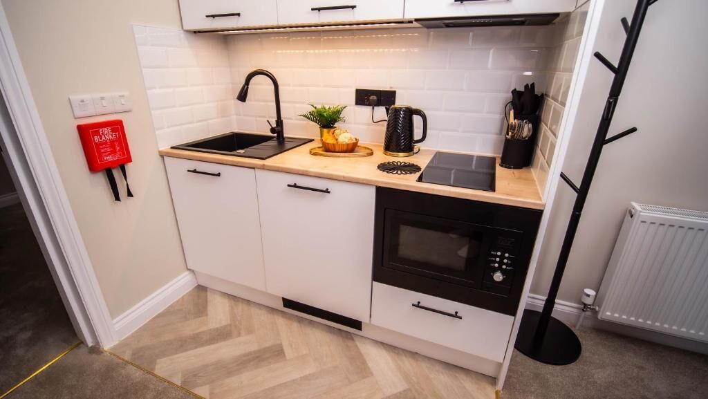A compact kitchen with white cabinets, a black stove, and a sink. A fire blanket is mounted on the wall.