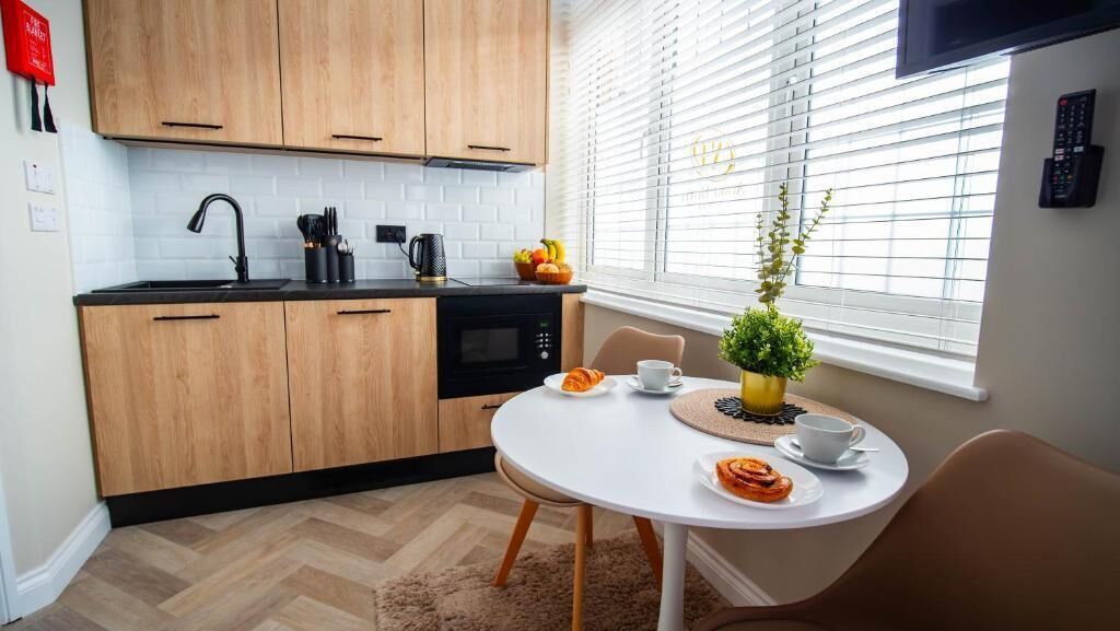 A modern kitchen with wooden cabinets, a black countertop, and a microwave. There is a dining table with a white tablecloth, a potted plant, and a fruit bowl.