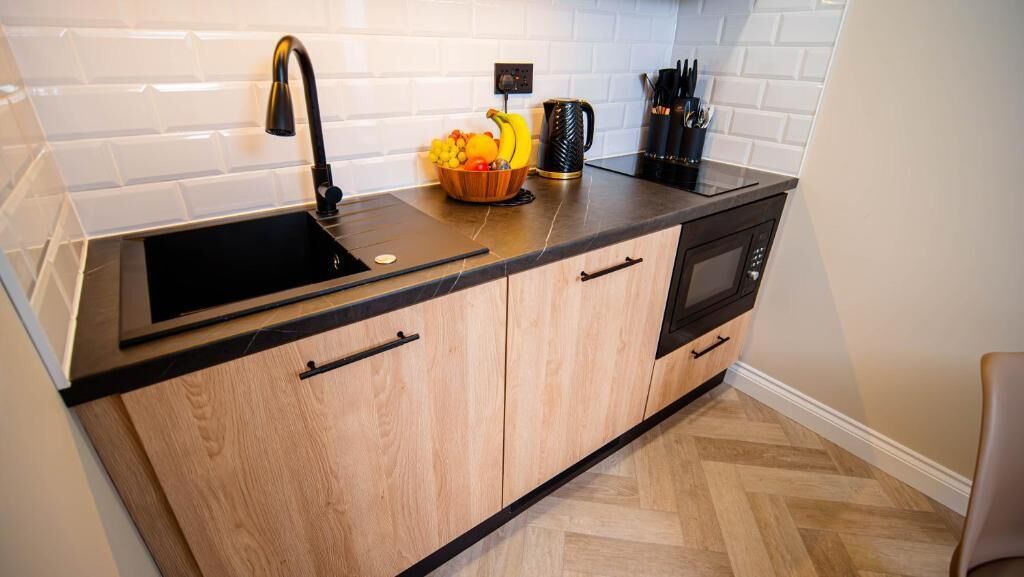 A modern kitchen with a black sink, wooden cabinets, a microwave, and a fruit bowl on the countertop.