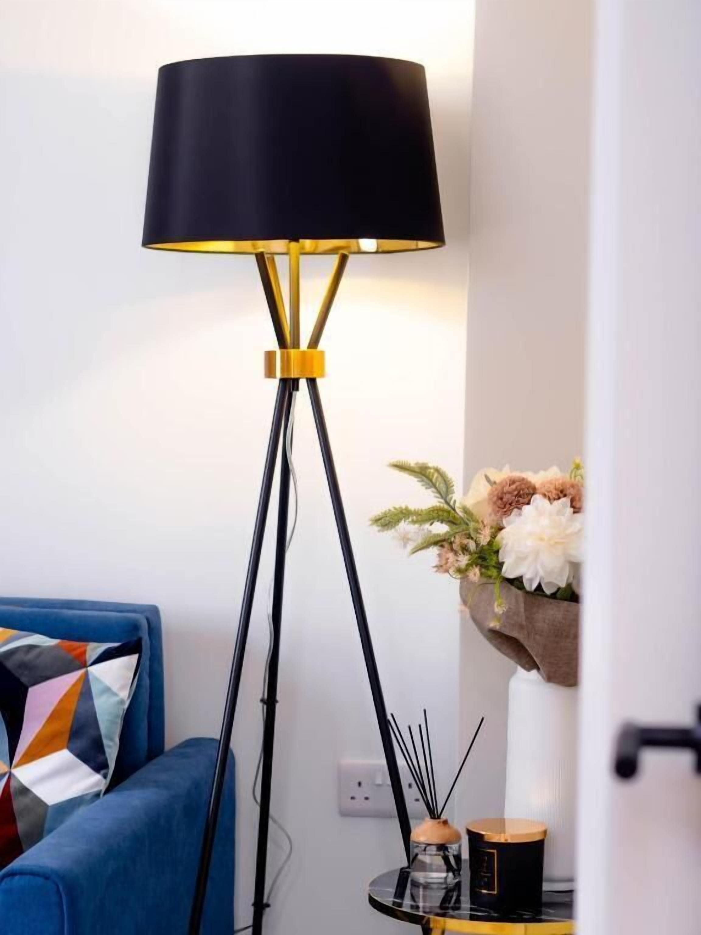 A modern living room with a black floor lamp with gold accents, a blue sofa with a geometric-patterned pillow, and a small round table with a candle and a vase of flowers.