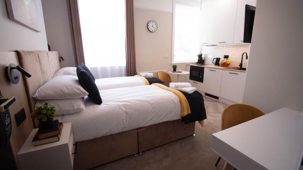 A hotel room with two beds, a small kitchenette, a desk, and a chair.