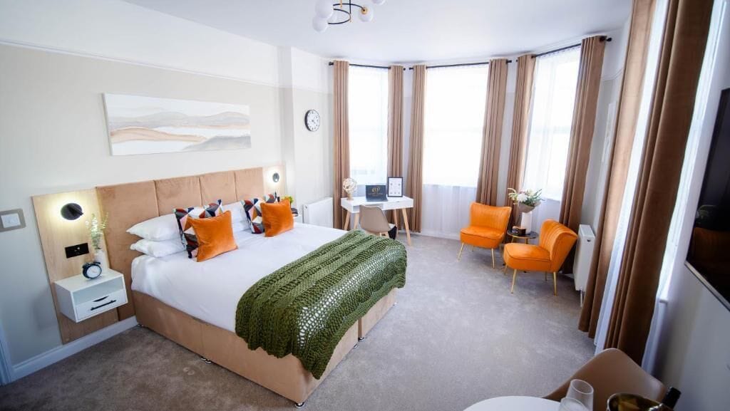 A hotel room with a bed, a desk, an orange chair, and a large window with curtains.