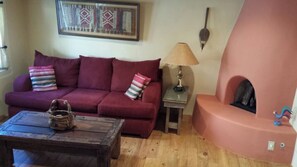 A cozy living room with a working Kiva fireplace, a red sofa, a wooden coffee table, and a lamp. - Historic Mill House Unit 4 (Taos)