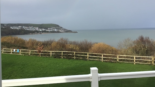 A coastal view with a grassy foreground, a wooden fence, and a distant town by the sea. - Lodge - Sleeps 4, Near Beach & Free Parking (New Quay)