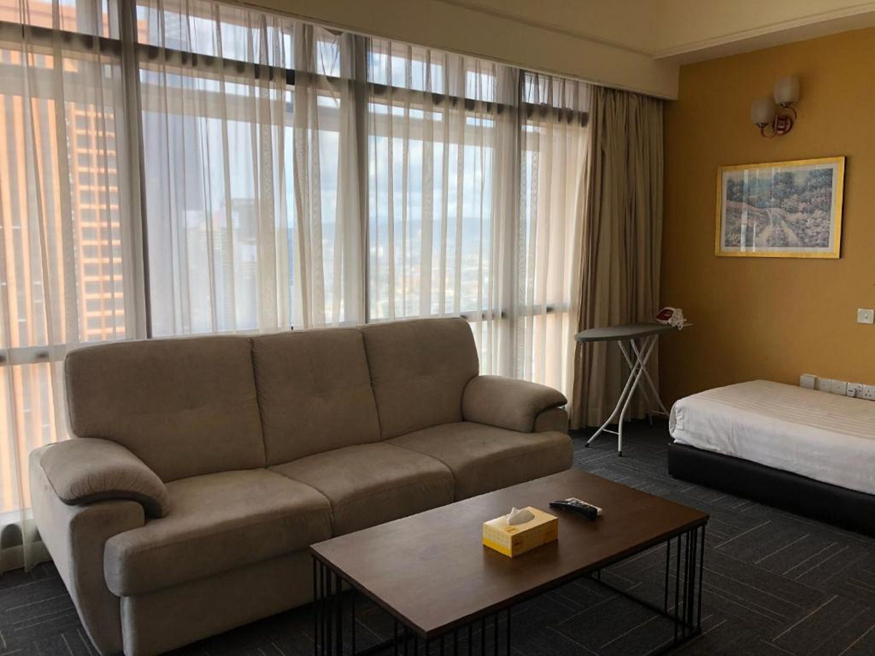 Ein Hotelzimmer mit beiger Couch, einem kleinen Holztisch mit Taschentuchbox und einem Bett mit weißer Bettwäsche, sowie einem Fenster mit durchsichtigen Vorhängen.