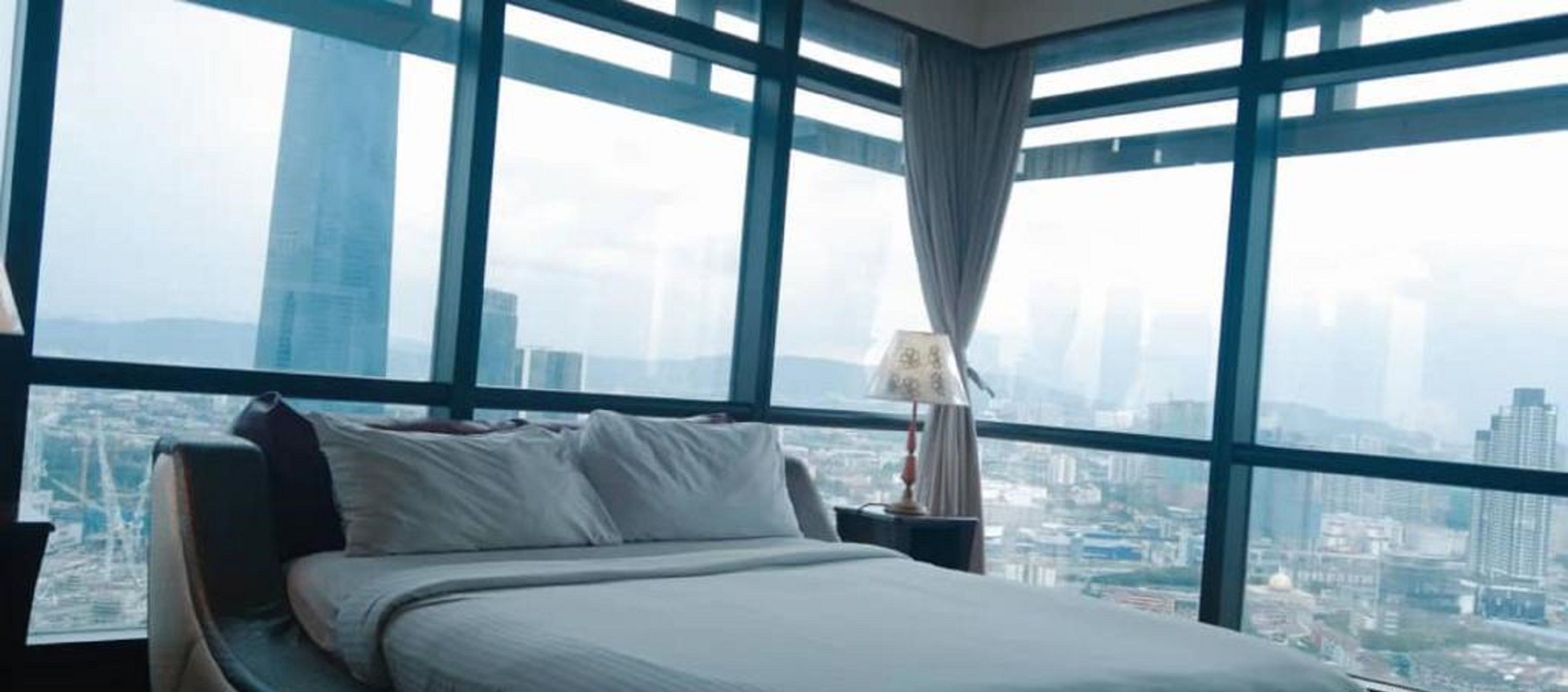 Habitación de hotel con una cama grande, una mesita de noche, una lámpara y vistas de la ciudad a través de ventanas del suelo al techo.
