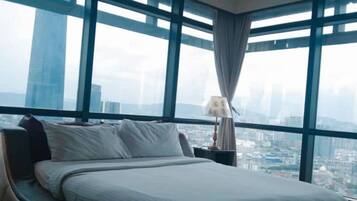 Habitación de hotel con una cama grande, una mesita de noche, una lámpara y vistas de la ciudad a través de ventanas del suelo al techo.