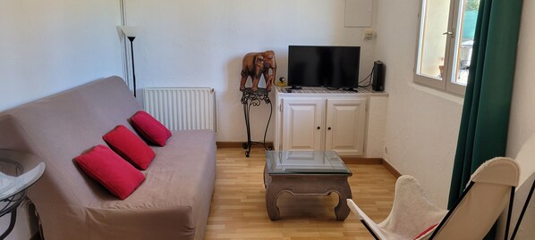 Una sala de estar con sofá, una mesa de centro de vidrio, un televisor sobre un mueble bajo y una ventana con cortinas.
