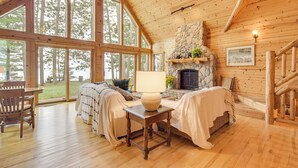 A log cabin interior with a fireplace, large windows, wooden furniture, and a stone accent wall.