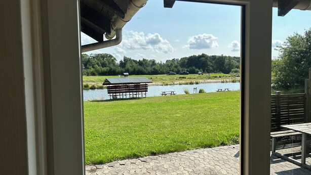 Vista de un área de picnic junto al río con un banco de madera y mesas, una pequeña construcción de madera, y un sendero pavimentado.