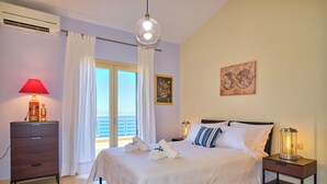 A neatly made bed with white bedding, a red lamp on a wooden nightstand, a globe artwork on the wall, and a view of the outdoors through a window with white curtains. - Villa Kallisti - Sea View & Private Pool (Corfu)