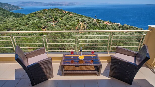 A balcony with wicker furniture and a table, overlooking a coastal landscape with olive trees and a view of the sea. - Villa Kallisti - Sea View & Private Pool (Corfu)