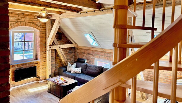 Una habitación estilo loft con vigas de madera a la vista, chimenea de ladrillo, sofá y escalera.