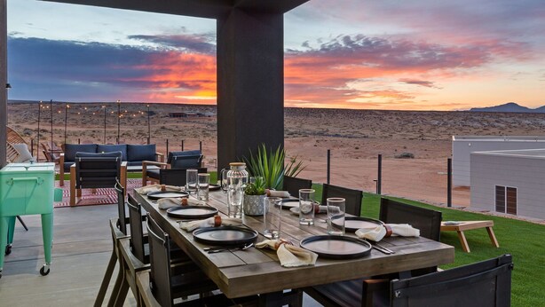 Un espacio de comedor al aire libre con vistas a un paisaje desértico al atardecer, que presenta una larga mesa de madera preparada para dos personas, asientos exteriores y un pequeño edificio al fondo.