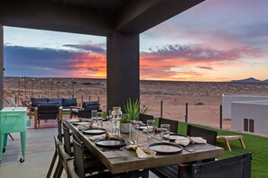An outdoor dining area with a view of a desert landscape at sunset, featuring a long wooden table set for two, outdoor seating, and a small building in the background. - Powell's Edge Retreat - Hot tub+ Pickle-ball + Home Gym + Lake Views + Sleeps 20 (Page)