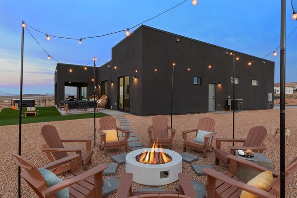 An outdoor seating area with Adirondack chairs and a fire pit, surrounded by a gravel patio and a modern building in the background. - Powell's Edge Retreat - Hot tub+ Pickle-ball + Home Gym + Lake Views + Sleeps 20 (Page)